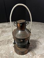 Front view of vintage brass masthead mariners hanging oil lamp with rope handle on gray stone surface against black backdrop.