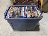 Blue plastic tote bin filled full with music CD jewel cases arranged upright in rows.