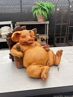 Front and angled views of the resin pig figurine holding a wine bottle and cup, seated against a wooden fence.