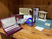 Full lot view showing all items together on wood surface: Lenox dolphin figurine, Lenox small clock in box, Arthur Court condiment bowl, Silver Treasures spreaders in presentation case, iridescent blue art glass heart, Mikasa dolphin bottle cork in box.