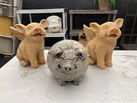 Three outdoor pig sculptures displayed on a shelf: a round concrete pig in gray color and two seated light brown resin pigs with wings.