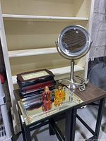 Vanity items on table including wooden jewelry box with glass top, lighted magnifying mirror, mirrored tray with perfumes