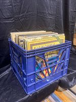 Blue slotted crate filled with various vinyl record albums visible from the front and top sides, showing colorful spines and partial cover artwork.