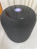 Side and top view of black Apple HomePod smart speaker showing mesh fabric and white Siri light on glossy surface.