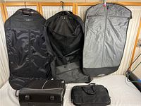 Three garment bags hanging on a rack, two black and one gray, with two carry bags placed in front on the surface