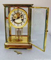 Front view of bevelled glass mantle clock with right door open, showing enamelled dial, pendulum and original winding key on base