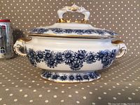 Side view of antique Royal Worcester sauce tureen showing blue floral pattern, elephant head handle with gold accents, and lid with chicken leg finial handle.