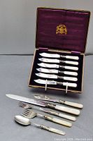 Full boxed set of six fruit knives with mother of pearl handles and silver blades with four additional serving utensils, presented inside a purple lined leather type box showing wear.