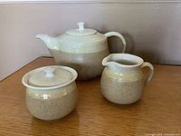 Front view of Northwind Stoneware trio tea set on wooden surface, showing teapot, creamer and lidded sugar bowl in beige and cream speckled glaze.