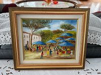 Painting in gold-toned frame with white matting showing outdoor market scene with blue umbrellas and people