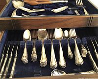 Two-tier wooden fitted case open showing flatware service – top tray with serving pieces and lower tray with forks, spoons, knives.