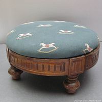 Side view showing wooden base and one of four turned feet, blue-green upholstery with embroidered rocking horses visible