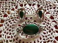 Front view of the oval sterling silver brooch with green Eilat stone and pair of matching teardrop-shaped malachite earrings with silver accents, displayed on white lace fabric.