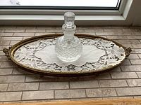 Perfume bottle centered on the antique oval tray and lace doily, viewed from side angle near window.
