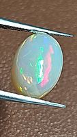 Oval cabochon cut Ethiopian opal held with tweezers showing bright play-of-color against textured background