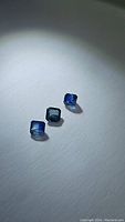 Three natural blue sapphires cut in emerald shape. Stones are dark to medium blue, placed on a white surface under light.