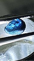 Detailed close-up of the teal blue green sapphire held by tweezers under magnification showing briolette faceted cut.