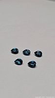 Five pear-shaped London blue topaz gemstones evenly spaced on a plain background showing cuts and color.
