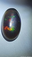 Close-up photo of an oval cabochon natural black opal showing multicolored flashes of orange, green, and blue on a dark base.