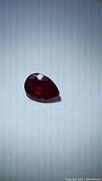 Photo of a pear-shaped faceted red ruby gemstone with visible internal features lying on a light surface.