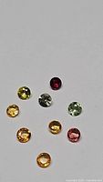 Nine small round gemstones of various colors including yellow, orange, green, pinkish-orange, and deep red arranged on a white background.