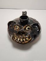 Front view showing grotesque face with painted eyes, teeth, nose, and ears on dark glazed pottery jug