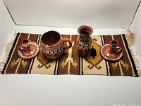 Photo of two small brown pottery candle holders, a brown pottery creamer or pitcher, and a brown pottery vase displayed on a Native American style woven rug with geometric patterns.
