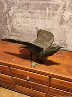 Bronze crow sculpture standing on a wooden surface, wings partially spread and beak open.