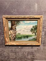 Full view of the framed oil painting showing the waterside scene with palm trees and cloudy sky.
