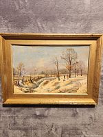 Frontal view of the signed oil painting depicting a winter landscape with snow, trees, and two figures walking. Painting is framed in a gold-toned wooden frame.