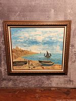 Front view of oil painting showing seascape with boats on shore and water, buildings along coast, partly cloudy sky, in ornate gold frame.