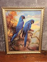 Front view of the framed oil painting of two blue parrots perched on a branch with plant and sky background.