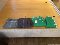 Four clothing items laid on bed: gray 'rockout' hoodie, dark zip-up hoodie, green Adidas track jacket, and matching green Adidas pants.