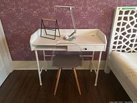 Front view of white desk set with lamp, chair, and storage rack against floral wallpaper.