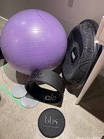 Purple exercise ball, black Rock Body balancer, Alo ring, and two athletic sliders on floor by wall and table.