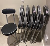 Six black vinyl padded folding chairs stacked folded against a wall