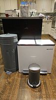 Three garbage cans grouped together in kitchen setting: large gray metal can with broken footstep, white Songmics dual-compartment recycle bin, small stainless steel pedal bin.