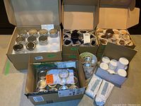 Boxes containing various sizes of glass canning jars with metal lids, metal tins, white plastic pots, and additional lids and sealing rings.