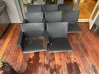 Eight stacked black wicker patio chairs arranged with four chairs with armrests and four without. Chairs show minor leaf debris and mild outdoor wear.