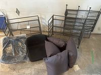 Three metal shoe racks, two black chair seats without legs, three dark gray throw pillows on garage floor against wall.
