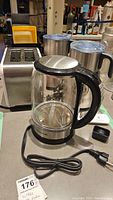 Shows Cosori electric glass kettle with black handle, clear body, power cord, and stainless steel base in front of toaster and frothers