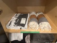 Three Turkish towels displayed on a wooden shelf, one is black and white with fringe, two are rolled grey and white patterned towels with paper bands.