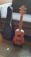 Oscar Schmidt Aloha ukulele standing on a black stand next to soft carrying case on floor in front of couch