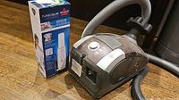 Miele vacuum with hose and power dial alongside boxed Bissell Turboslim hand vacuum on floor surface.