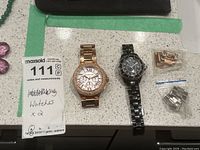 Two wristwatches placed side by side with bags of extra links beside them and a MaxSold tag showing quantity 2.