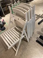 Set of six white folding chairs stacked and bound with green strap, showing side profile and visible slatted wood seats and folding hinges.