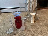 Group of 12 assorted vases in glass, metal, and china, arranged on carpeted floor near baseboard heater and window.