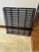 Folded metal dog pen, black color, showing side view and top view, leaning against wall and floor.