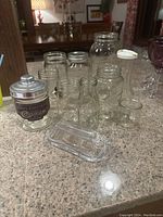 Six assorted mason jars, one with metal lid, small containers, and glass butter dish with lid arranged on counter.