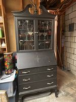 Black wooden secretary desk, original green paint covered with black paint. Shows three drawers, two intact, and glass doors on upper cabinet portion.